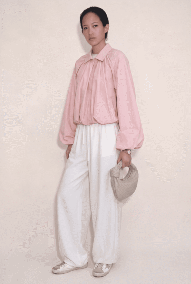 Woman in a pale pink blouse with voluminous sleeves and white wide-leg pants, holding a small beige woven handbag against a neutral backdrop.