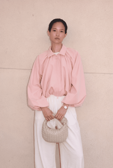 Woman stands against a beige wall in a pale pink zip-up jacket and white pants, holding a woven beige bag in front.
