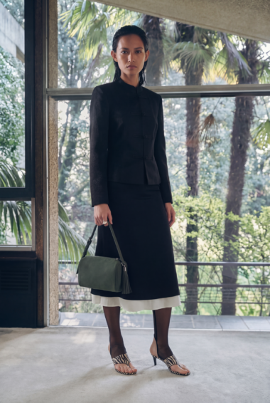 Woman in a black suit-style jacket and long skirt stands indoors by a glass wall, holding a green handbag.