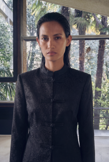 Portrait of a woman with slicked-back hair wearing a black textured Mandarin-collar jacket, standing indoors by a window with tropical plants outside.