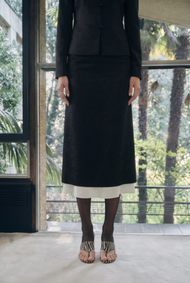 Full-length view of a person indoors wearing a black tailored suit with a long skirt and zebra-striped sandals in front of a glass door.