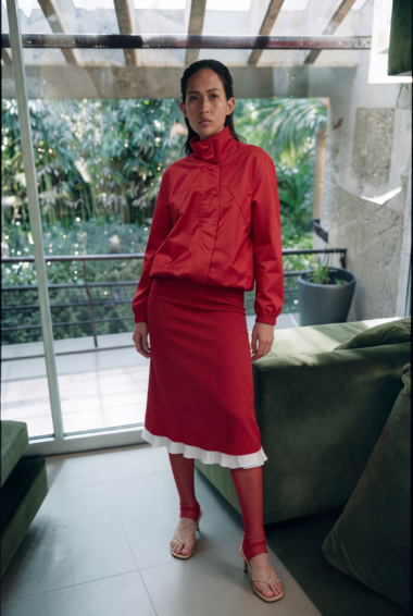 Woman in a red high-neck blouse and matching red skirt with white ruffled hem, standing in a bright, modern living space with greenery outside.