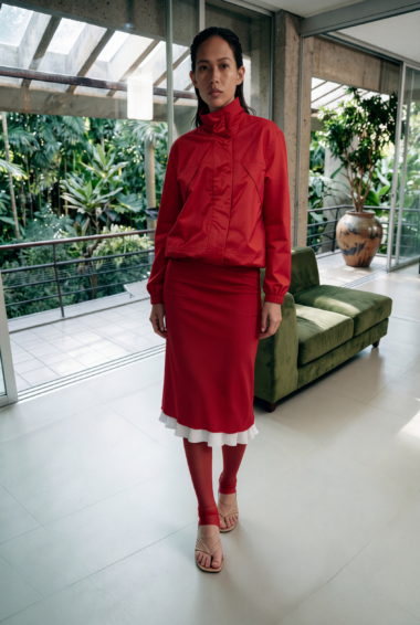 Woman in a red high-collar jacket and red skirt stands indoors beside a green sofa, with glass walls and tropical plants outside.
