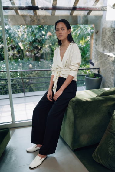 Woman seated on a green sofa in a modern indoor space, wearing a cream wrap blouse and black wide-leg pants, looking at the camera.