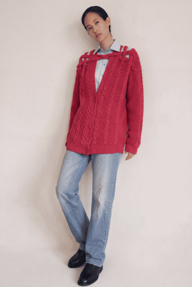 Young person wearing a bright red oversized cable-knit cardigan over a light blue striped button-down shirt and light-wash jeans, posing against a neutral wall.