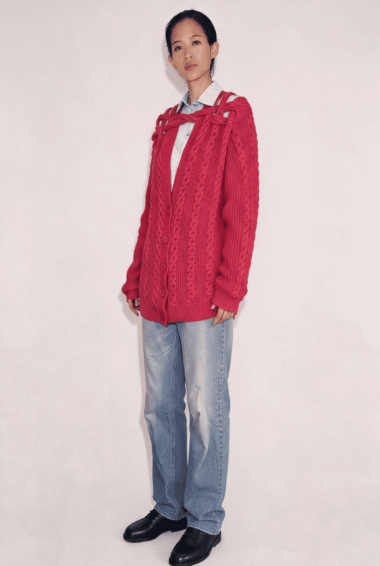 Model wearing a chunky red cable-knit cardigan over a light striped shirt, blue jeans, and black loafers, standing against a pale backdrop.