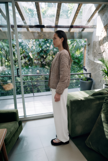 Woman standing in profile beside glass doors, wearing a beige floral-embroidered jacket and white pants, gazing toward the garden.