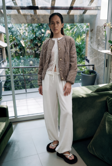 Woman standing in a glass-walled lounge wearing a taupe floral-embroidered jacket over a white lace top and wide-leg white pants.