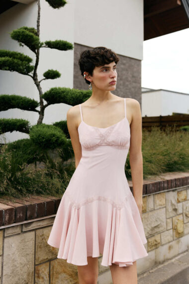 Woman in a pale pink dress standing outdoors beside a stone wall and trimmed greenery, looking to the right.