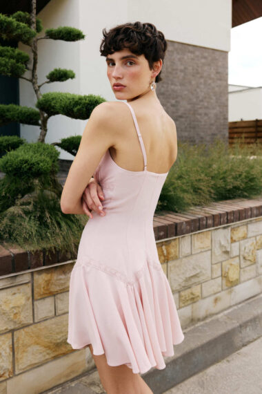 Woman in a pale pink sleeveless dress standing outdoors, back to camera, looking over shoulder with short dark hair.