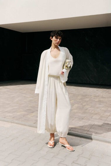 Woman in an all-white outfit (tank top, wide-leg pants, and long robe) stands on a paved outdoor area, holding a floral clutch.