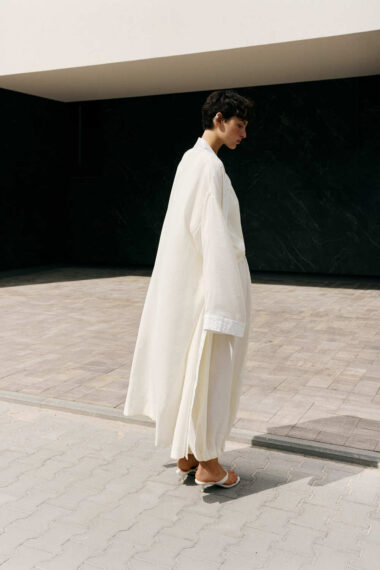 Person in a long white robe walking sideways on a paved outdoor area, sandals visible, against a dark wall and light overhang (side view).