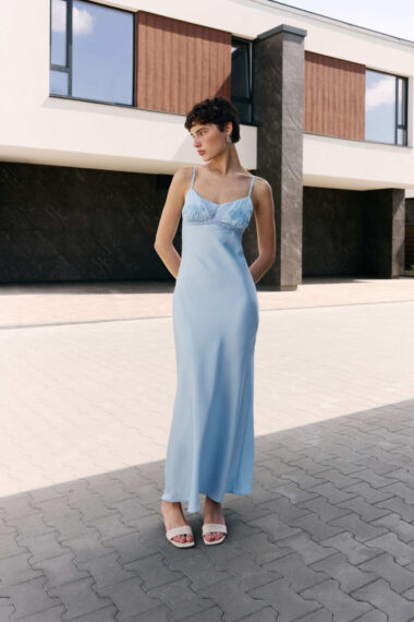 Woman in a pale blue satin slip dress with lace bodice stands on a paved plaza in front of a modern building, hands behind her, wearing white sandals.