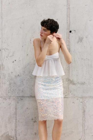 Woman in a white spaghetti-strap top and sequin skirt adjusts an earring against a concrete wall.