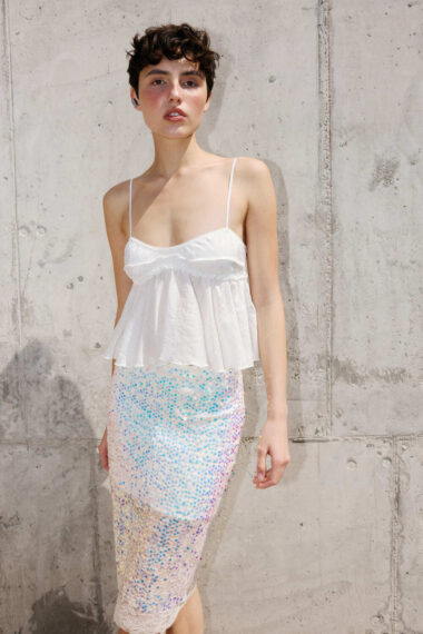 Model posing against a gray concrete wall in a white spaghetti-strap top and pastel sequin midi skirt.