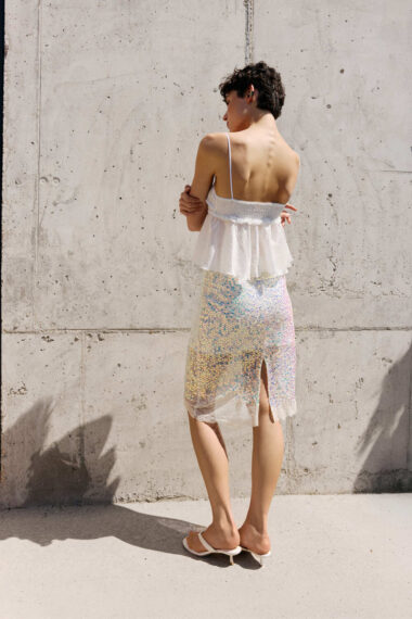 Back view of a person wearing a white peplum top and iridescent sequin skirt, standing against a concrete wall in sandals.
