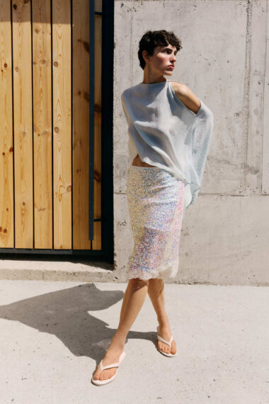 Woman wearing a pale blue off-shoulder top and iridescent sequin midi skirt stands beside a concrete wall in sandals.