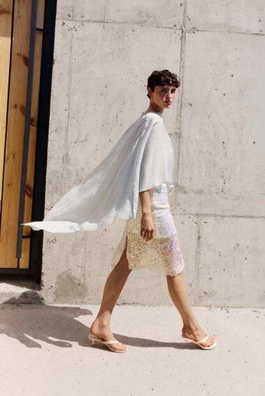 Model walking in a light gray cape over a pastel sequin skirt beside a concrete wall.