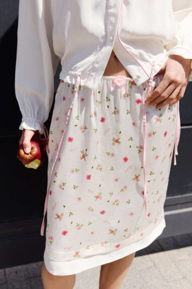 Person wearing a white blouse with pink trim and a white floral skirt, holding a partially eaten red apple at side.