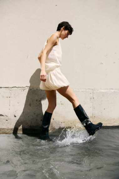 Person in a white sleeveless dress and black rain boots walking through shallow water beside a beige concrete wall, splashing as they step.