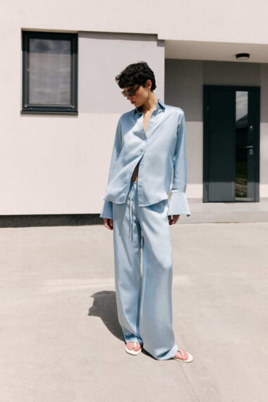 Person in a pale blue oversized shirt and matching wide-leg pants stands outdoors beside a modern beige building, wearing sunglasses.