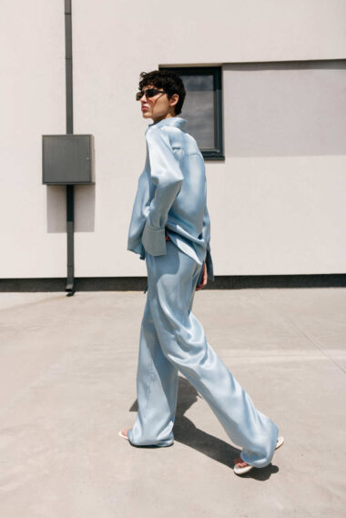 Person in a light blue satin suit walking outdoors beside a beige building, wearing sunglasses.