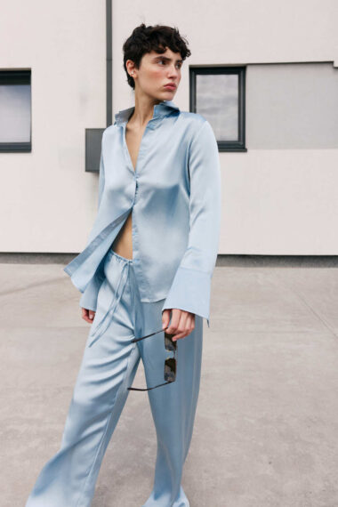 Person in a light blue satin shirt and matching wide-leg pants walks outdoors, holding sunglasses in one hand.