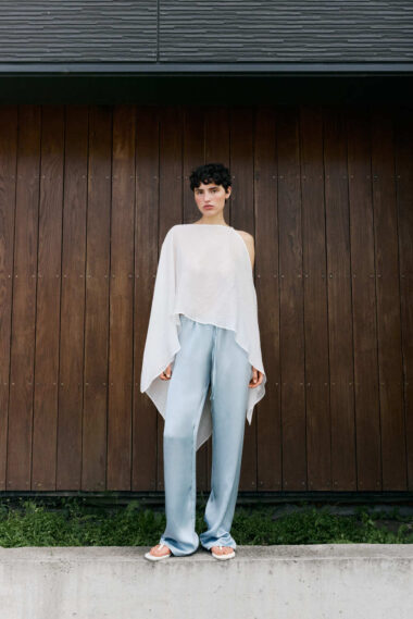 Model wearing a white, asymmetrical poncho top and pale blue wide-leg pants, standing on a concrete ledge in front of a dark wooden wall.