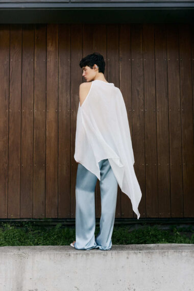 Person with short black hair wearing a white asymmetrical sleeveless top and light blue wide-leg pants, standing sideways against a wooden wall.