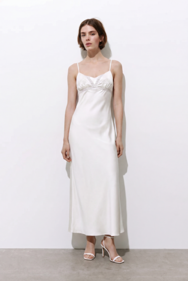 Woman in a white satin slip dress with lace bodice, spaghetti straps, standing against a white wall wearing white heeled sandals.