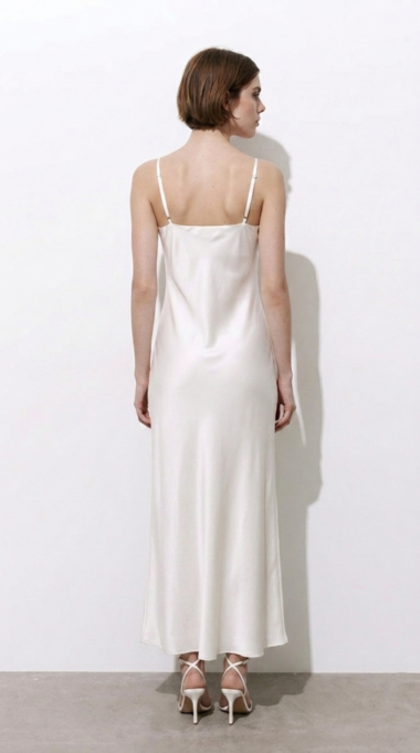 Back view of a woman in a white satin slip dress with thin spaghetti straps, standing against a light wall in strappy white heels.