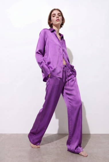 Model wearing a purple satin pajama set with a button-down shirt and drawstring wide-leg pants, barefoot against a white backdrop.