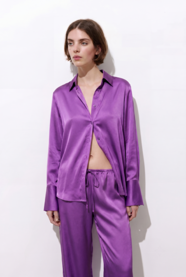 Woman wearing a shiny purple satin pajama set with an open shirt, midriff visible, standing against a white wall.