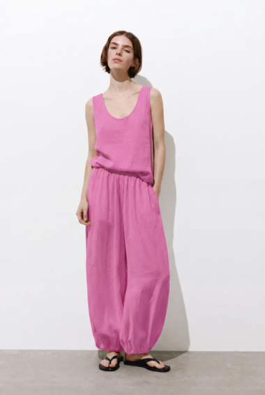 Full-length portrait of a woman in a bright pink sleeveless jumpsuit with an elastic waist against a white background, wearing black flip-flops.