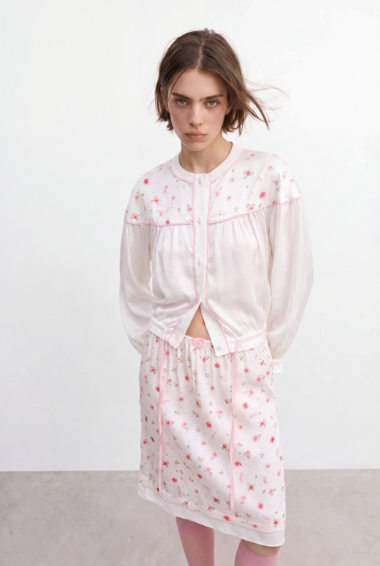 Model wearing a pale pink floral pajama set with long sleeves, pink piping, and a knee-length skirted shorts, standing against a white background.