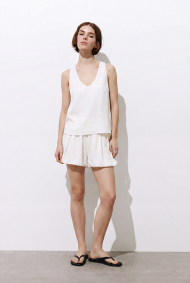 Woman in a white sleeveless top and white shorts standing against a white backdrop, wearing black flip-flops.