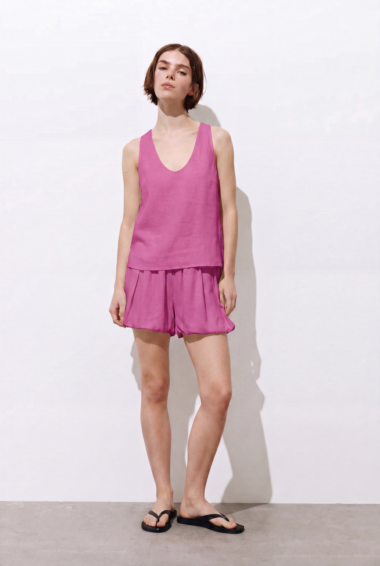 Woman in a pink sleeveless top and matching pink shorts, standing against a white backdrop in black flip-flops