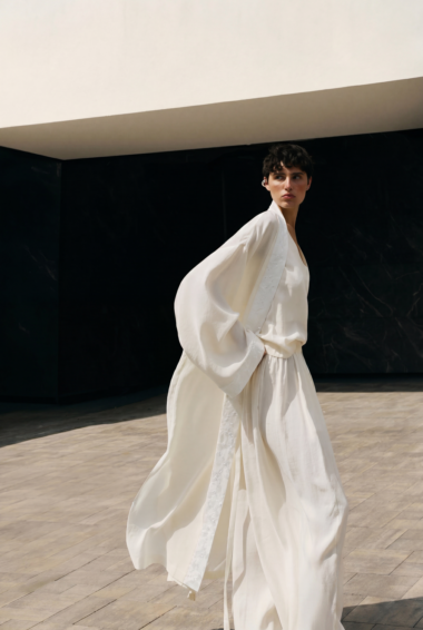 A person in a flowing white robe walks outdoors on a stone courtyard against a dark wall.