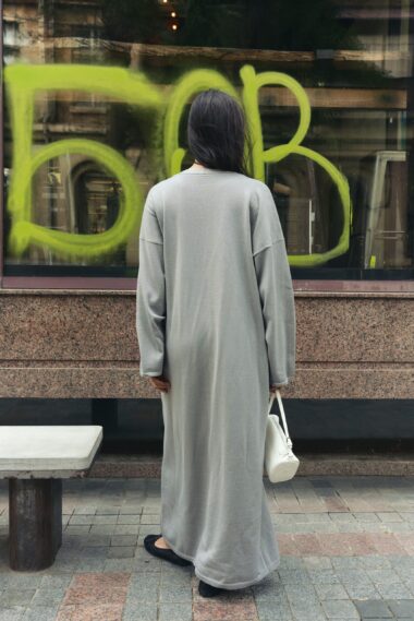 Person with long dark hair in a gray dress stands with their back to the camera in front of a window with neon green graffiti, holding a white bag.