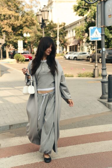 Woman in light gray joggers and long coat walks across a city crosswalk, holding a phone and small white purse.