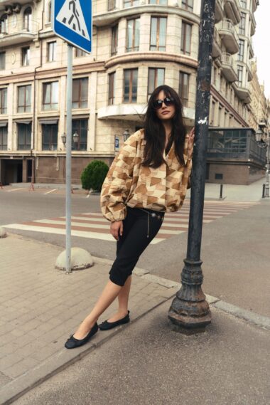 Woman in a geometric beige jacket and black pants leans against a dark lamppost on a city street, wearing sunglasses.