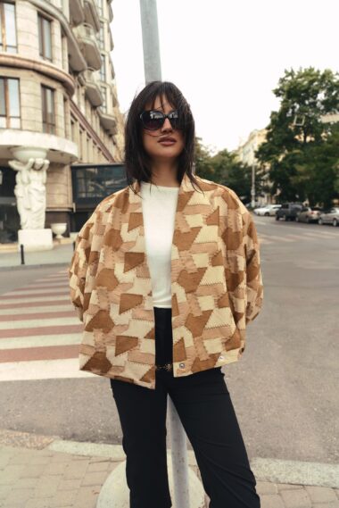 Woman in a tan geometric-patterned coat and black pants, wearing sunglasses, standing on a city street by a lamppost.