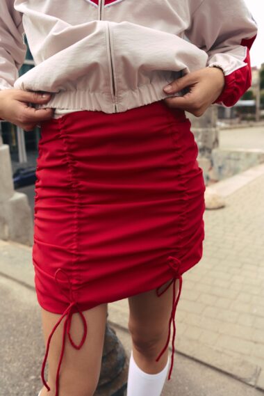 Person wearing a beige zip-up jacket adjusts a red ruched mini skirt outdoors, showing the waist area.