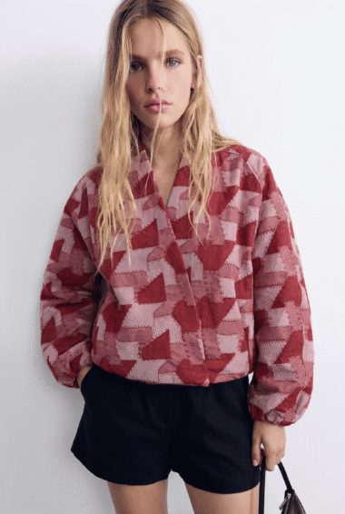 Young woman standing against a white wall, wearing a red geometric-patterned jacket and black shorts, holding a brown handbag.