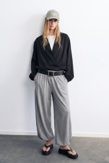Woman standing against a light gray wall wearing a black wrap blouse, gray wide-leg pants, a beige cap, and black sandals.