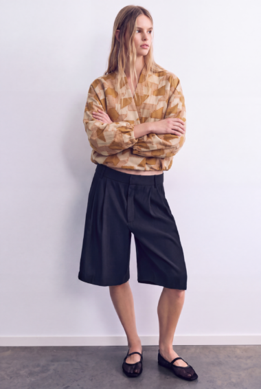 Woman standing in a studio, wearing a beige geometric blouse and navy Bermuda shorts, arms crossed.