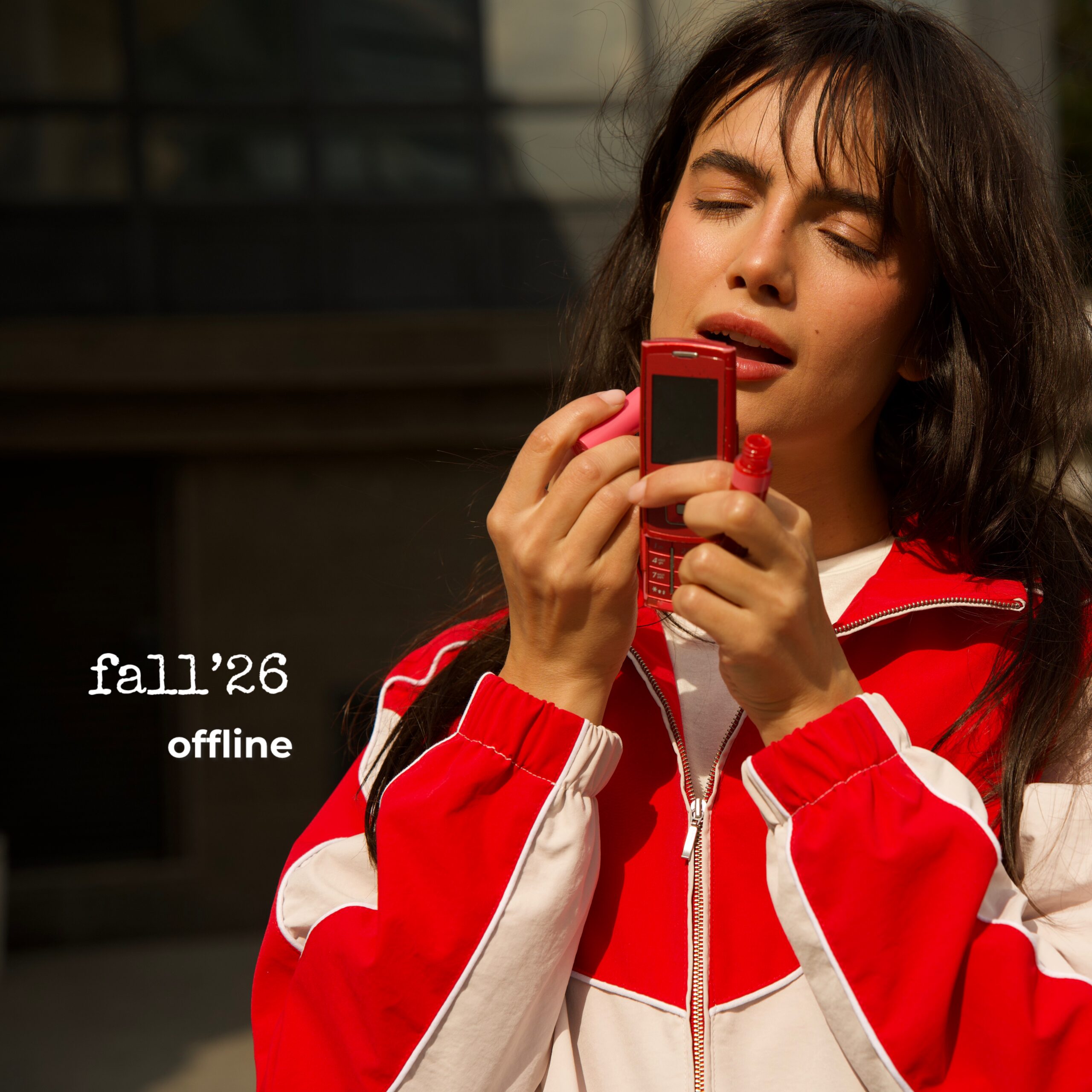 Young woman with long dark hair, wearing a red and white jacket, holding a red handheld device near her mouth with eyes closed outdoors.