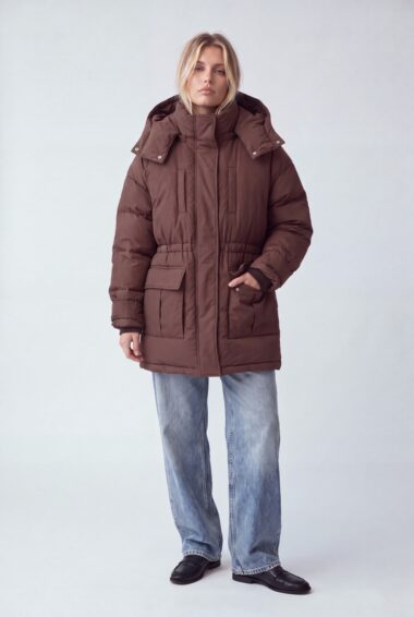 Woman modeling a long brown puffer coat with large front pockets over light blue jeans.