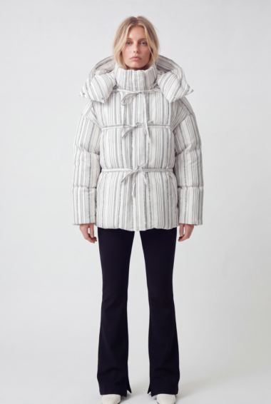 Model wearing a white striped quilted puffer jacket with a high collar and multiple tie-fastens, paired with black pants and white shoes.