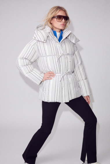 Woman modeling a white striped belted puffer jacket, blue layer peeking out, black pants, and oversized sunglasses against a light background.